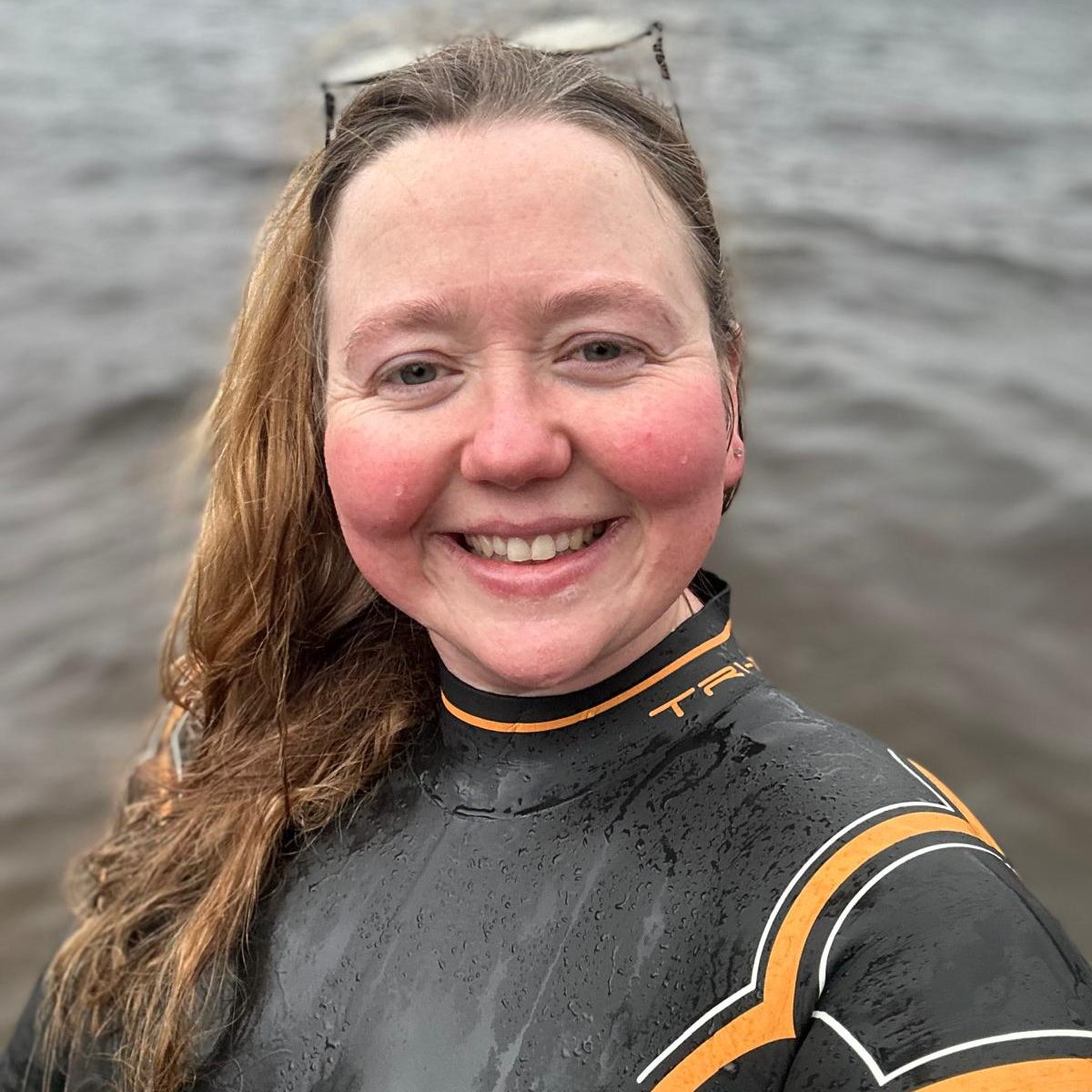 Smiling female, open water swimming coach, wearing a wetsuit, with water in the background.
